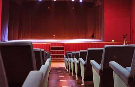 Interior del teatro con escenario iluminado y butacas grises en primer plano