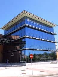 Fachada frontal del edificio con estructura de cristal azul y marquesina metálica, mostrando entrada principal