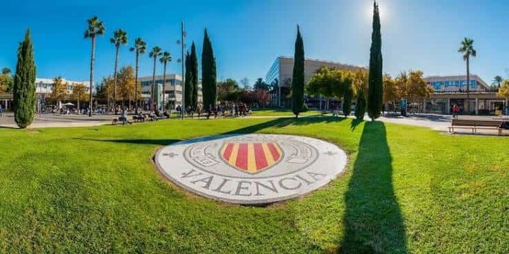 Plaza central del campus con el escudo de la UPV en el césped y palmeras al fondo