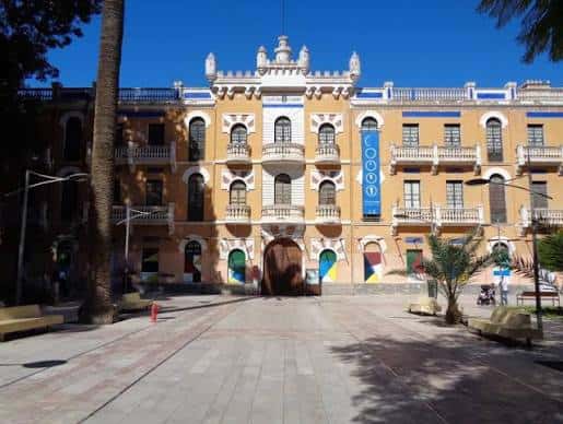 Vista frontal del edificio con balcones ornamentados y plaza adoquinada frente a la entrada