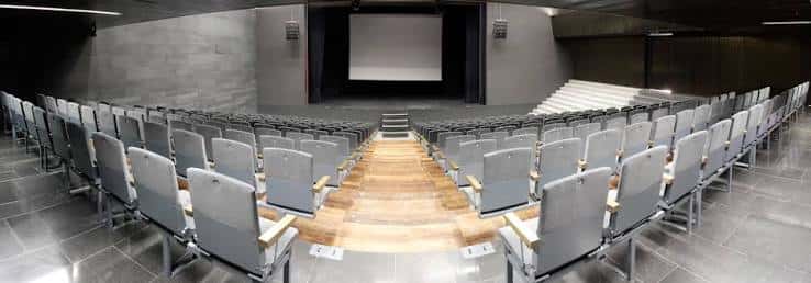 Sala de eventos o auditorio con butacas grises dispuestas en semicírculo frente a una pantalla