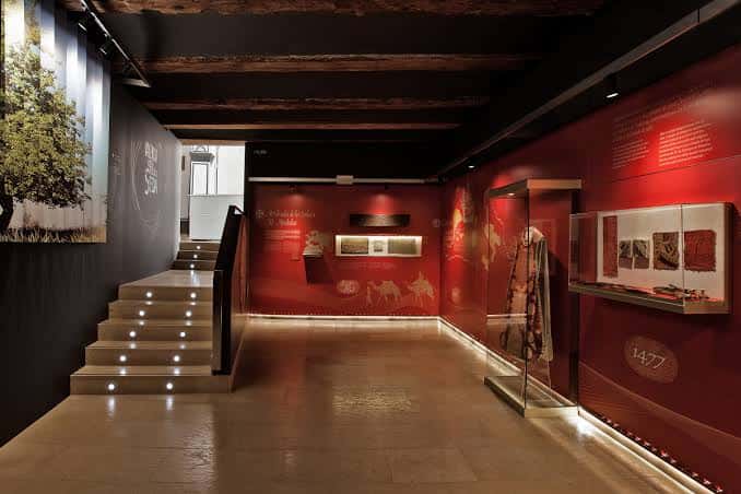 Interior del museo con escalera iluminada y paredes rojas exhibiendo arte textil