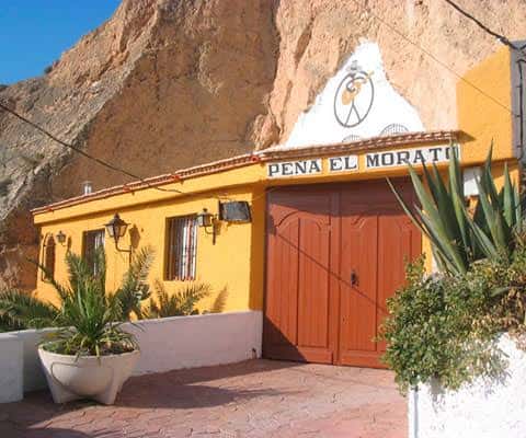 Fachada exterior de la Peña Flamenca El Morato, un edificio amarillo con puerta de madera y letrero distintivo, ubicado al pie de un acantilado