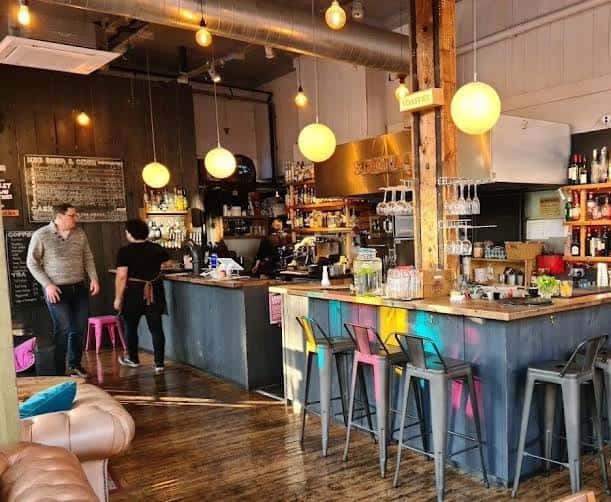 Interior del bar con barra de madera, taburetes metálicos y luces colgantes esféricas