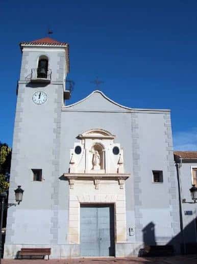 Vista frontal de la Iglesia de Nuestra Señora de Cortes bajo cielo azul, destacando su torre con reloj y nicho central
