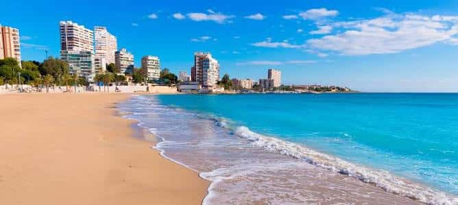 Litoral de la Playa de San Juan con olas rompiendo en la orilla y skyline urbano al fondo