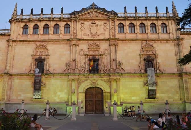 Fachada iluminada de la Universidad de Alcalá al atardecer con personas en el patio