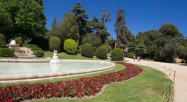 Vista panorámica de los jardines con fuente, sendero y visitantes bajo cielo azul