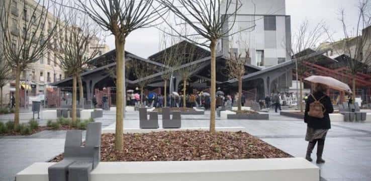 Plaza reformada con árboles jóvenes y bancos de hormigón frente al mercado