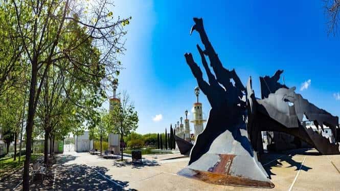 Escultura metálica en primer plano con las torres y árboles al fondo