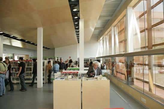 Interior de la biblioteca con estanterías, lectores y grandes ventanales que dejan entrar luz natural