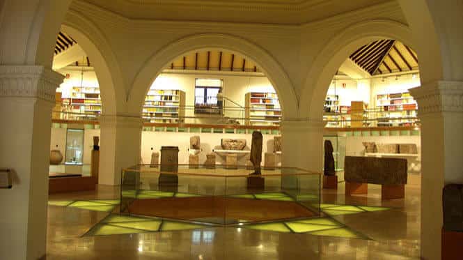 Salón central del museo con arcos, columnas y exposición de piezas arqueológicas bajo iluminación cálida