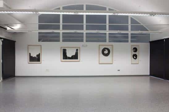 Sala de exposiciones interior con obras colgadas en pared blanca y suelo gris