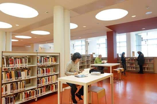 Interior de la biblioteca con estanterías, mesas de lectura y personas consultando