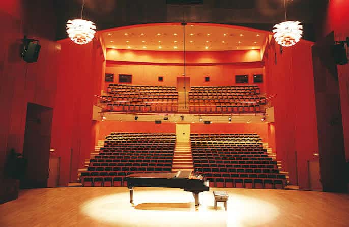 Interior del auditorio con escenario, piano y butacas vacías bajo iluminación cálida