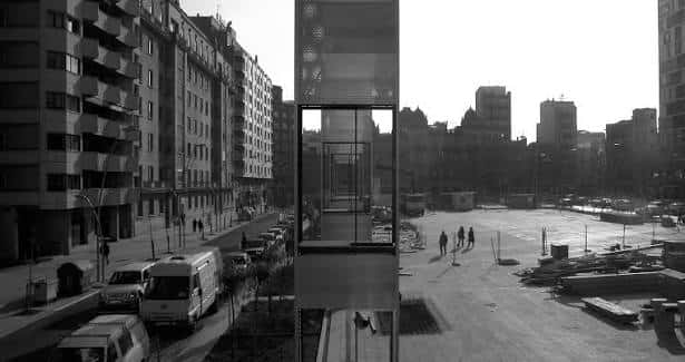 Vista en blanco y negro de la plaza con ascensor exterior y personas caminando