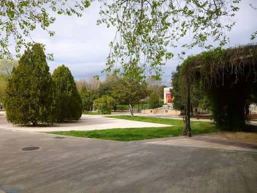 Vista general del parque con césped, árboles y pérgola cubierta de enredaderas
