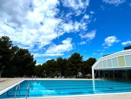 Piscina exterior del complejo Can Roca bajo un cielo azul con nubes y árboles al fondo