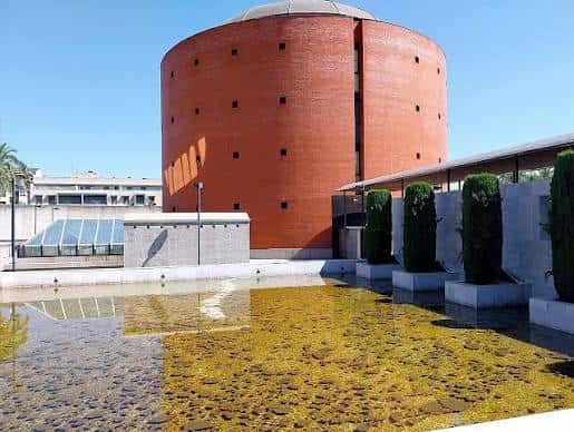 Fachada del Museo MEIAC reflejada en el estanque frontal, con palmeras y cielo despejado