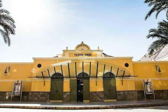Fachada principal del teatro en día soleado, con palmeras y entrada central