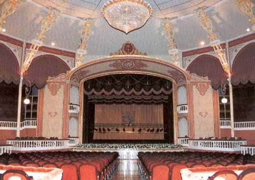 Interior del teatro con decoración clásica, lámpara de cristal y butacas rojas frente al escenario