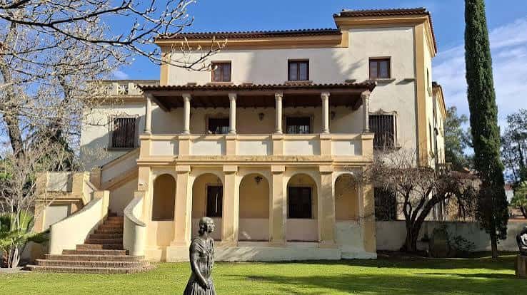 Fachada principal de la Casa Pedrilla con columnas y escalinata, rodeada de jardines y una escultura en primer plano