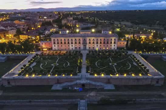 Vista aérea nocturna del palacio iluminado con los jardines ornamentales y la ciudad de fondo