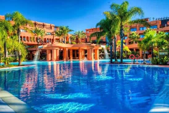 Vista de la piscina principal del hotel rodeada de palmeras y edificios de estilo andaluz