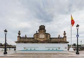 Vista frontal completa de la Fuente de las Galeras con bandera española y farolas