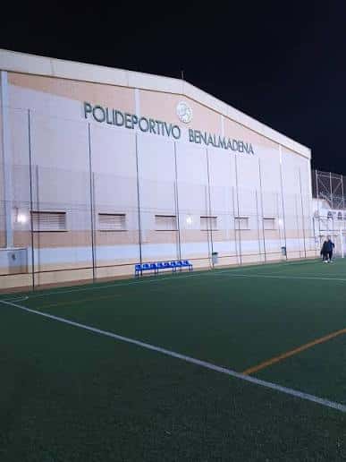 Fachada del polideportivo iluminada por la noche, con bancos y campo visible en primer plano