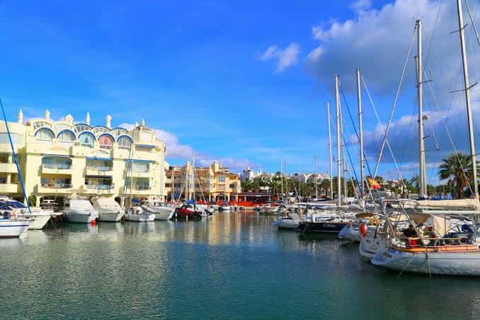 Puerto deportivo con veleros y yates bajo un cielo azul, mostrando la zona de atraque y edificios costeros