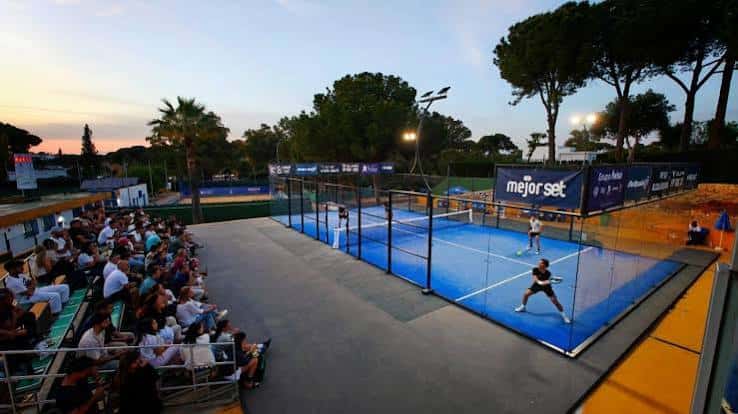 Partido de pádel en pista azul con espectadores en gradas al atardecer