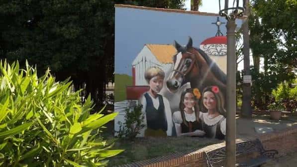 Mural artístico en la plaza con figuras infantiles y un caballo, rodeado de vegetación y farolas