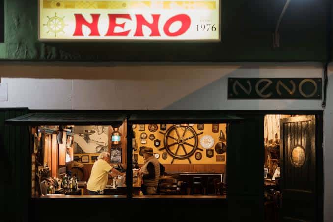 Fachada nocturna del Bar Neno con su letrero iluminado y vista al interior decorado
