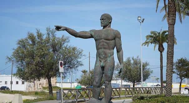 Estatua de bronce de un hombre desnudo en un entorno con palmeras y cielo azul