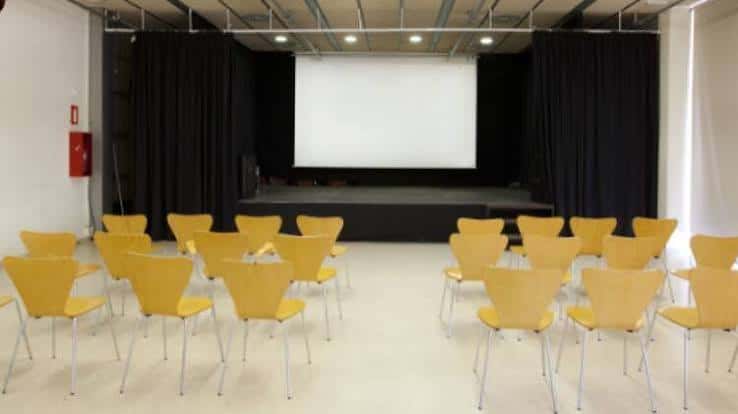 Sala de eventos interior con sillas amarillas dispuestas frente a una pantalla y escenario