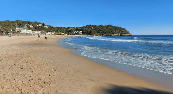 Playa amplia con olas suaves rompiendo en la orilla, personas caminando y colinas arboladas al fondo