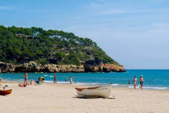 Escena playera con embarcaciones en la arena, bañistas disfrutando del sol y una colina boscosa al fondo