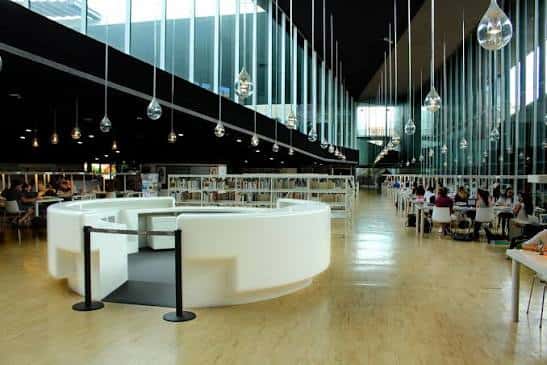 Área de lectura con mostrador circular blanco y filas de estanterías bajo el techo con lámparas suspendidas