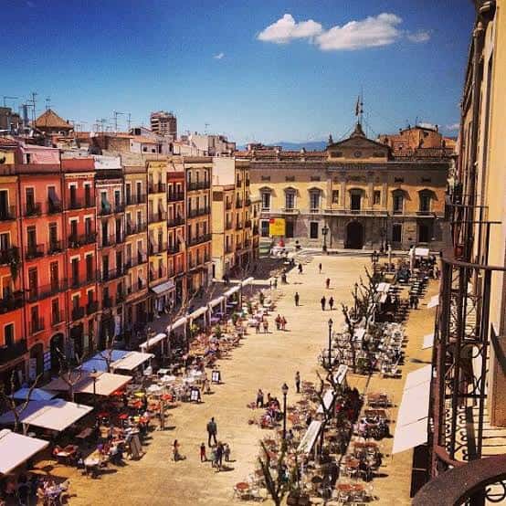 Vista elevada de la plaza llena de terrazas de cafeterías y gente paseando bajo un cielo azul