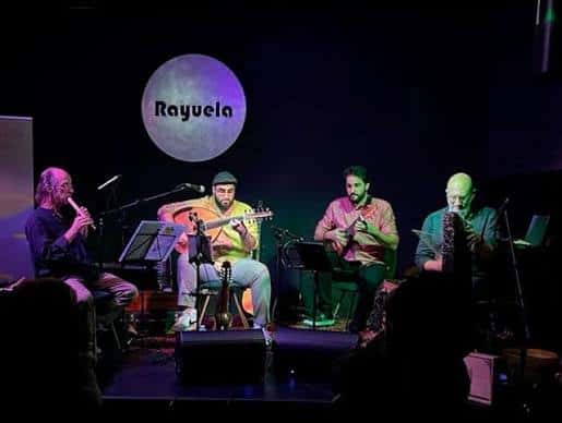 Concierto en vivo en el escenario del Café Teatro Rayuela con banda y logo proyectado