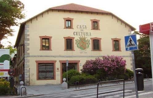 Fachada frontal de la Casa de la Cultura con arbustos floridos y señalización peatonal