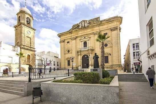 Plaza Mayor con estatua en primer plano, la iglesia y la torre al fondo, bajo un cielo parcialmente nublado