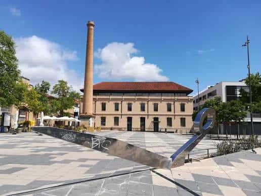 Vista panorámica de la Plaça de Cal Font con la chimenea industrial y la escultura metálica bajo un cielo azul