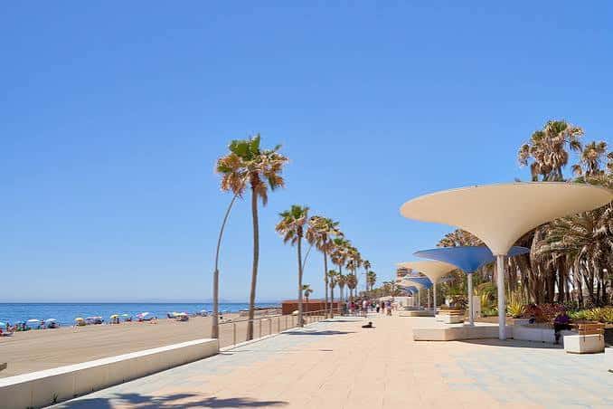 Vista diurna del paseo marítimo con sombrillas blancas y palmeras bajo cielo despejado