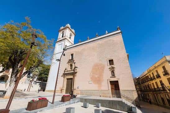 Fachada lateral de la iglesia con plaza y árboles en primer plano