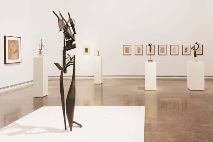 Galería blanca con escultura central de metal y obras enmarcadas en las paredes
