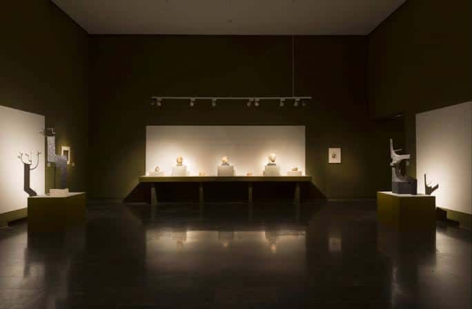 Sala oscura con esculturas sobre pedestales y mesa central iluminada