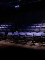 Interior del auditorio del teatro con filas de butacas azules bajo iluminación tenue, vista desde el escenario