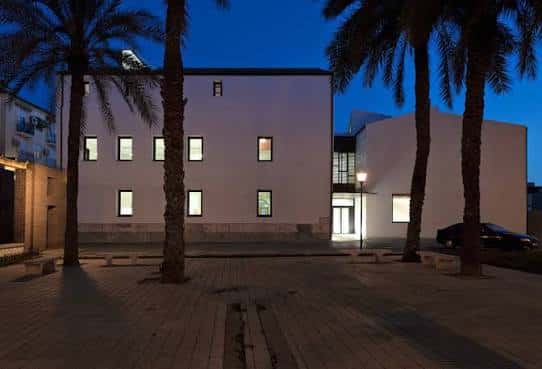 Fachada de la Biblioteca Pública María Moliner al atardecer, con palmeras iluminando la plaza y ventanas encendidas